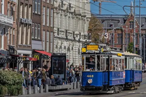 Tranvía histórico azul de la línea 3 con vagón remolque circulando por una calle concurrida del centro de Ámsterdam junto a la multitud