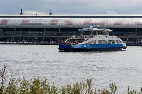 Ferry azul y blanco de GVB con pasajeros cruzando el río IJ, con la gran cubierta de cristal de Amsterdam Centraal al fondo