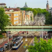 Tranvía azul y blanco de la línea 19 de GVB pasando bajo un puente ferroviario en una avenida de Ámsterdam con ciclistas y árboles