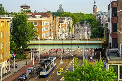 Tranvía azul y blanco de la línea 19 de GVB pasando bajo un puente ferroviario en una avenida de Ámsterdam con ciclistas y árboles