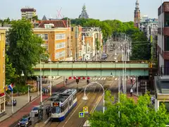 Tranvía azul y blanco de la línea 19 de GVB pasando bajo un puente ferroviario en una avenida de Ámsterdam con ciclistas y árboles