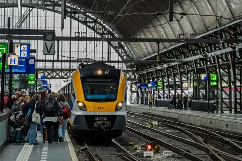 Viajeros con maletas esperando en un andén de Amsterdam Centraal mientras llega un tren amarillo de NS bajo la cubierta de hierro