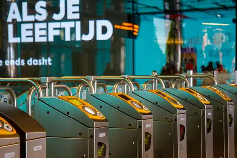 Hilera de barreras de acceso con lectores amarillos para tarjeta en una estación de metro de Ámsterdam, con carteles en neerlandés al fondo