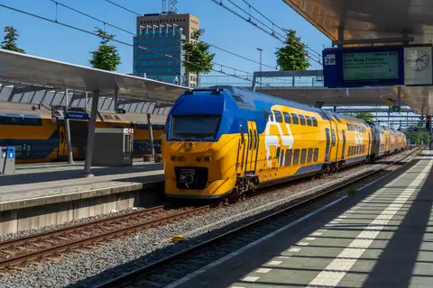 Tren NS de dos pisos, amarillo y azul, detenido en el andén 20b de Utrecht Centraal, con pantalla de salidas y torre al fondo