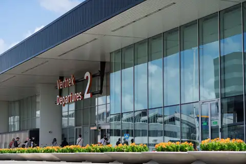 Fachada de cristal de la terminal de salidas 2 de Schiphol, con letrero Vertrek Departures, jardineras de flores y viajeros en la entrada