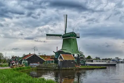 Molino de madera verde con aspas y casetas de madera oscura a la orilla del río Zaan, bajo un cielo cargado de nubes
