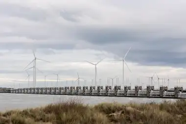 Pilares de hormigón de la barrera del Escalda Oriental sobre el agua, con aerogeneradores detrás, dunas en primer plano y cielo nublado